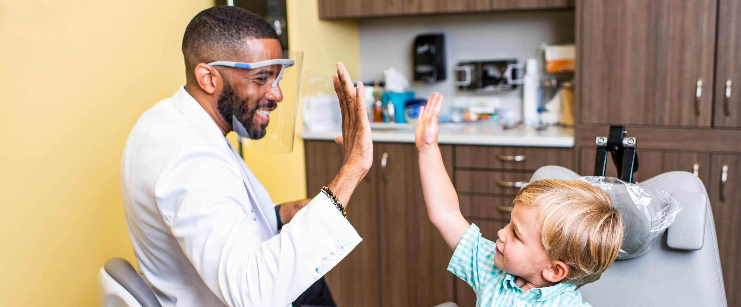 A smiling dentist in a clinical coat shares a high-five with a young child in the exam chair.