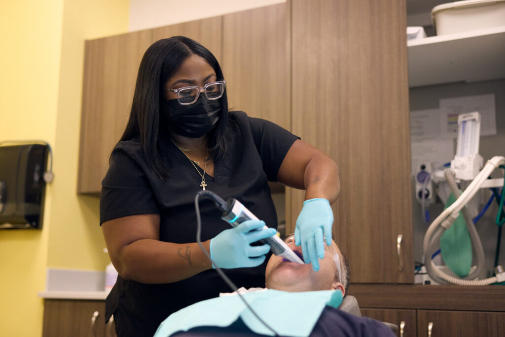 A dental professional wearing a face mask and gloves uses a handheld intraoral scanner to capture digital impressions of a reclining patient's teeth.