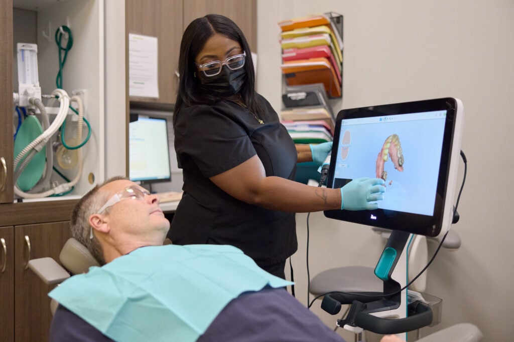 A dental professional wearing a face mask and gloves points to a 3D digital model of teeth on a monitor to explain a treatment plan to a reclining patient.