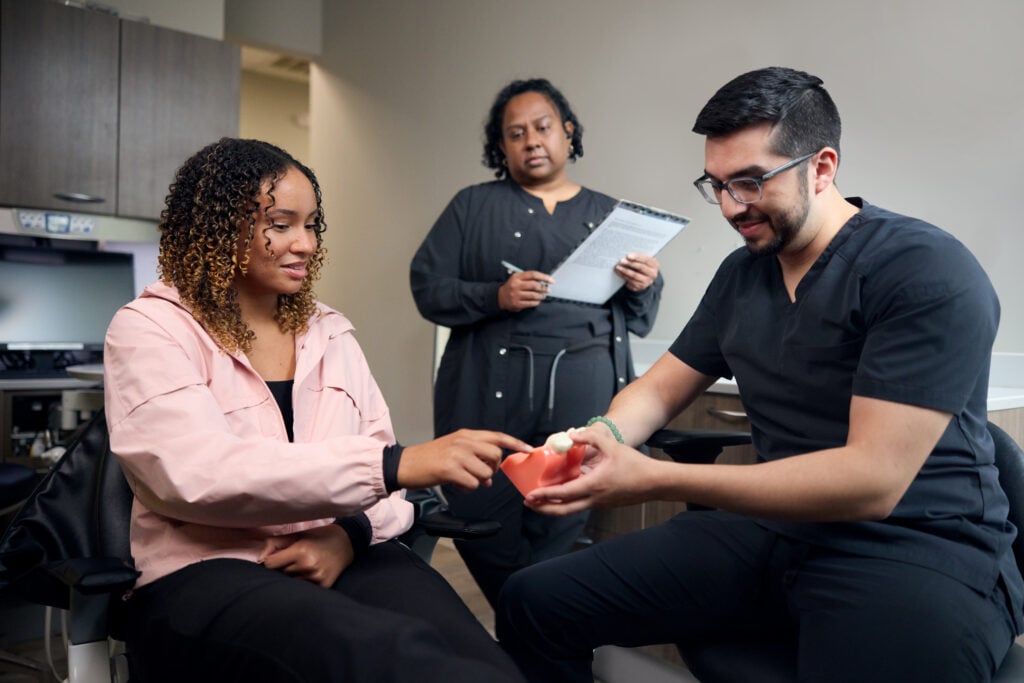 A patient points to a tooth on a dental jaw model held by her dentist during a consultation, while a dental assistant looks on with a clipboard.