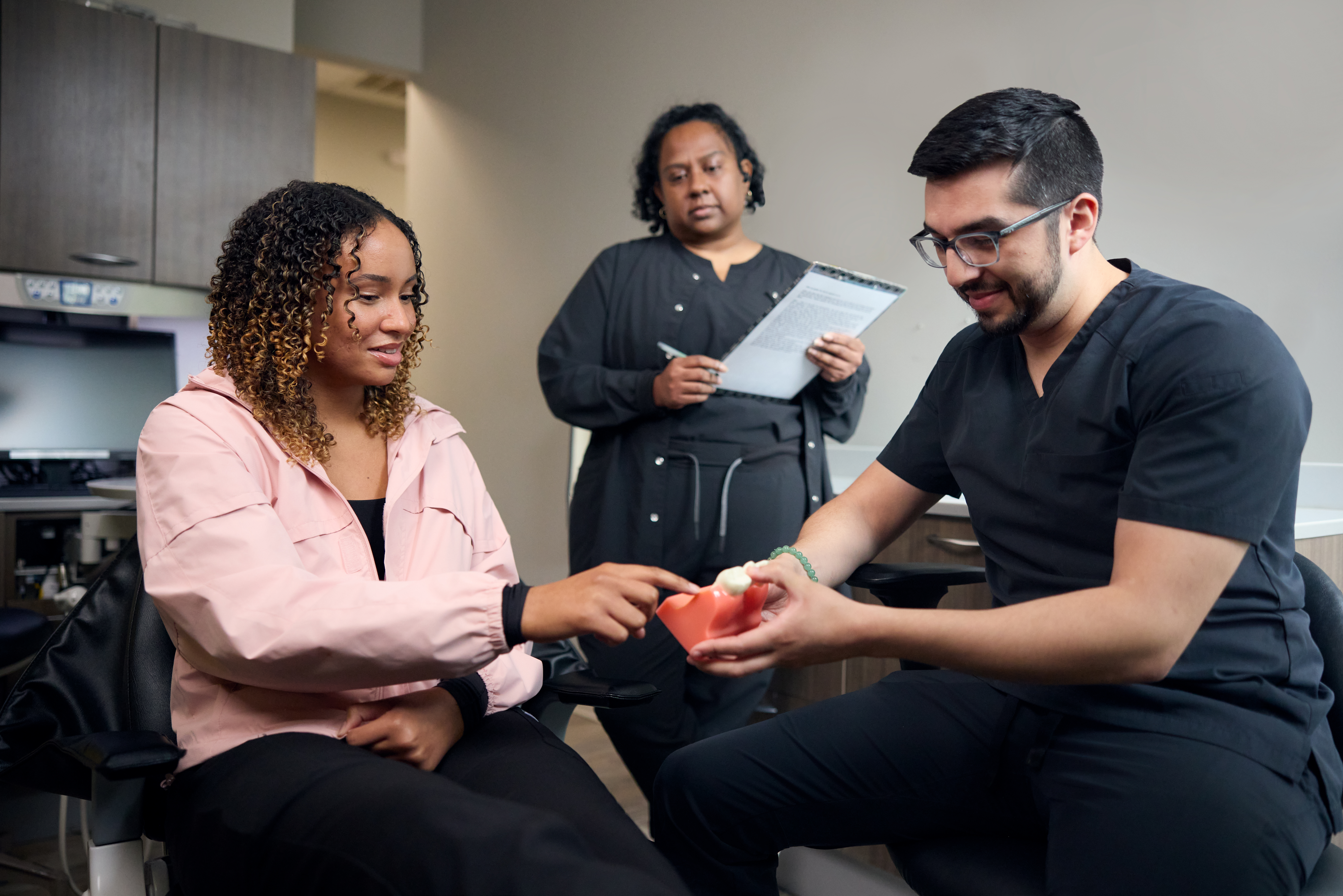 A seated female patient points to a specific tooth on an anatomical jaw model held by Dr. Ross Rosenblatt during a treatment consultation, while a dental assistant looks on with a clipboard.