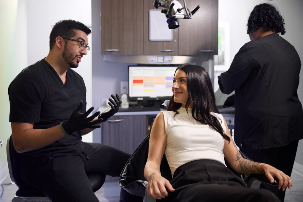A dentist wearing black scrubs and gloves sits and engages in a consultation with a patient, while a staff member works in the background.