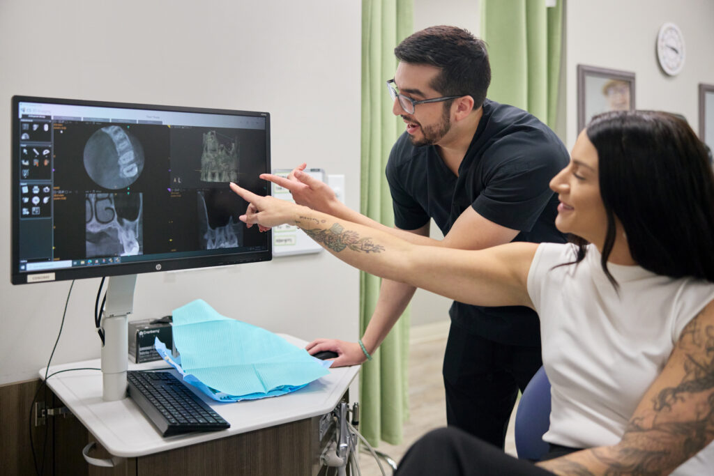 A dentist and a patient point to and review detailed dental X-ray imaging on a computer monitor during a clinical consultation.