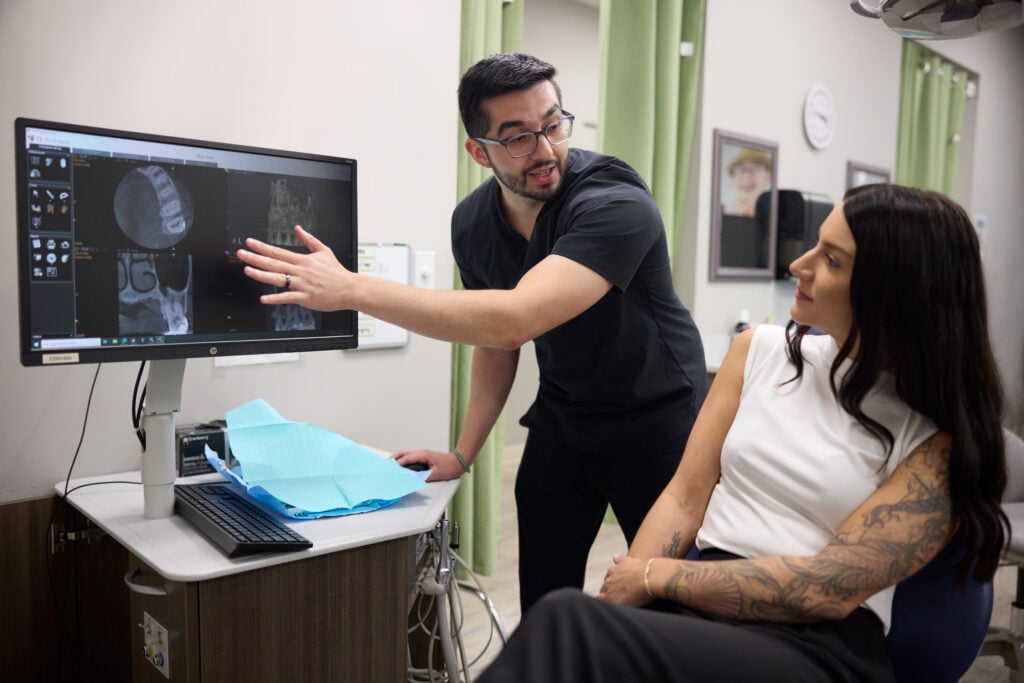 Dr. Rosenblatt points toward a computer monitor displaying dental imaging, providing a consultation to a patient seated nearby.