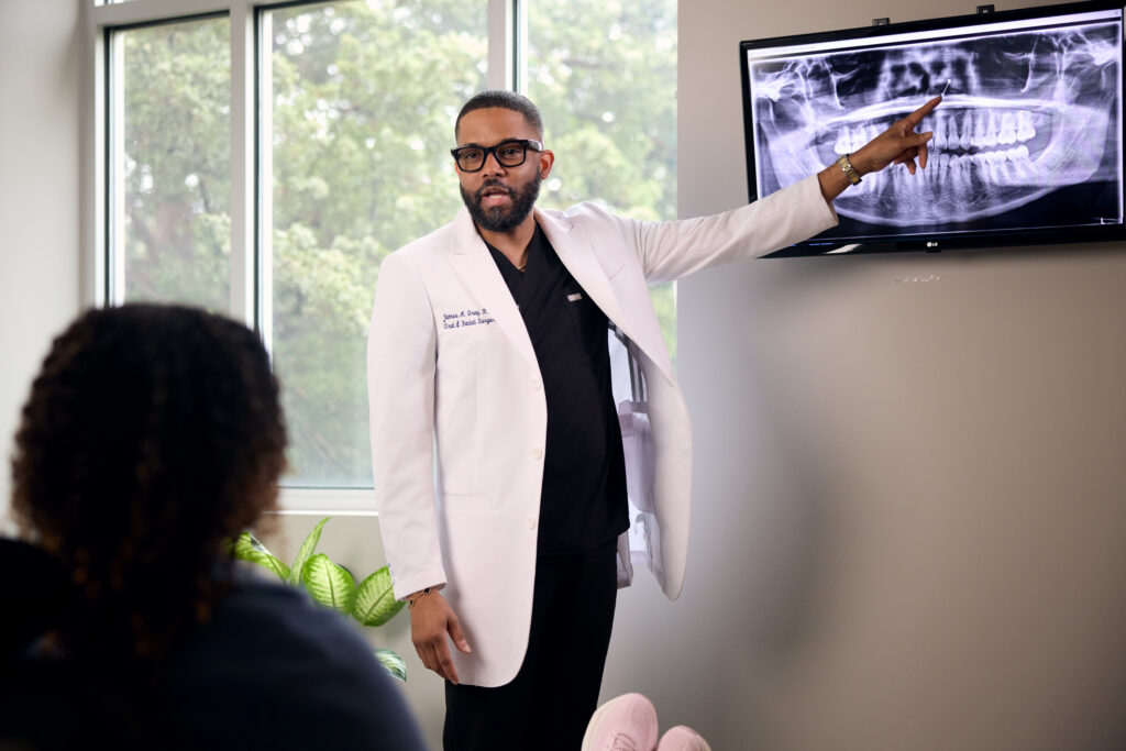 Dr. James Gray, an oral and facial surgeon in a white clinical coat, points to a panoramic dental X-ray displayed on a monitor while consulting with a patient.