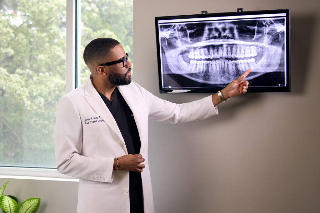 Dr. James Gray, oral and facial surgeon, in a white clinical coat points to a panoramic dental X-ray displayed on a wall-mounted monitor during a consultation.