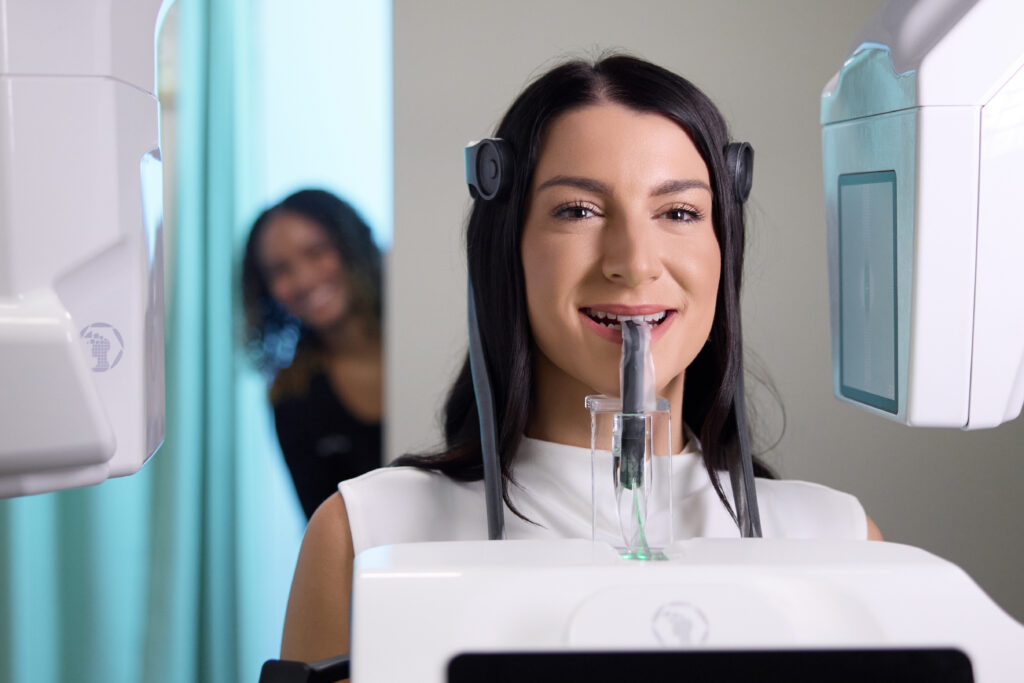 A female patient is positioned in a 3D dental imaging machine for a scan while a staff member smiles in the background.